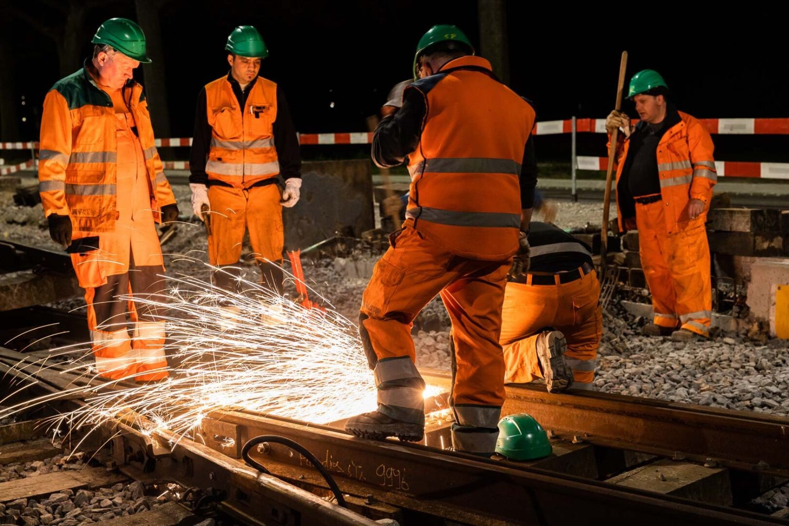 Workers in orange safety gear are repairing train tracks at night.
