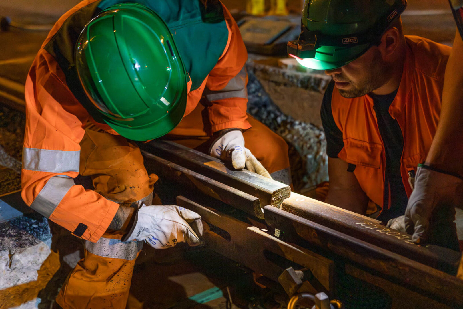 Two workers in orange safety gear are fixing a railway track at night.