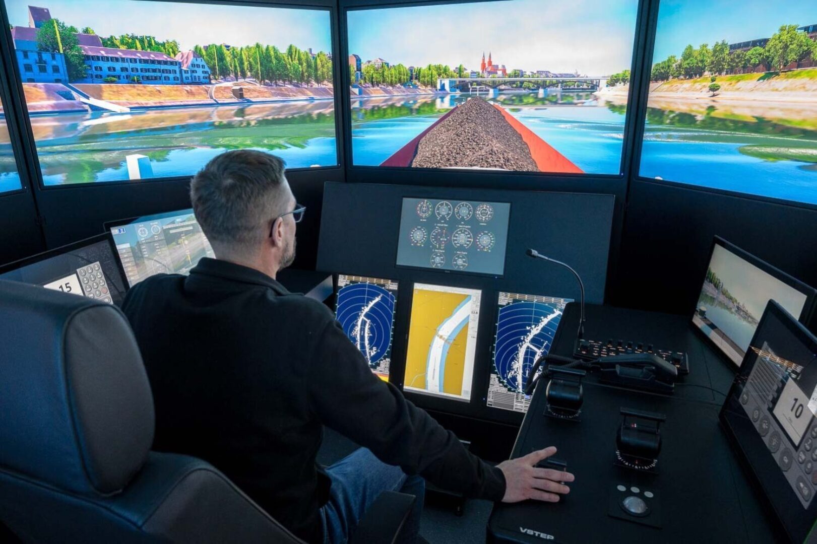 A person operates a simulation control panel with multiple screens displaying river and bridge views.