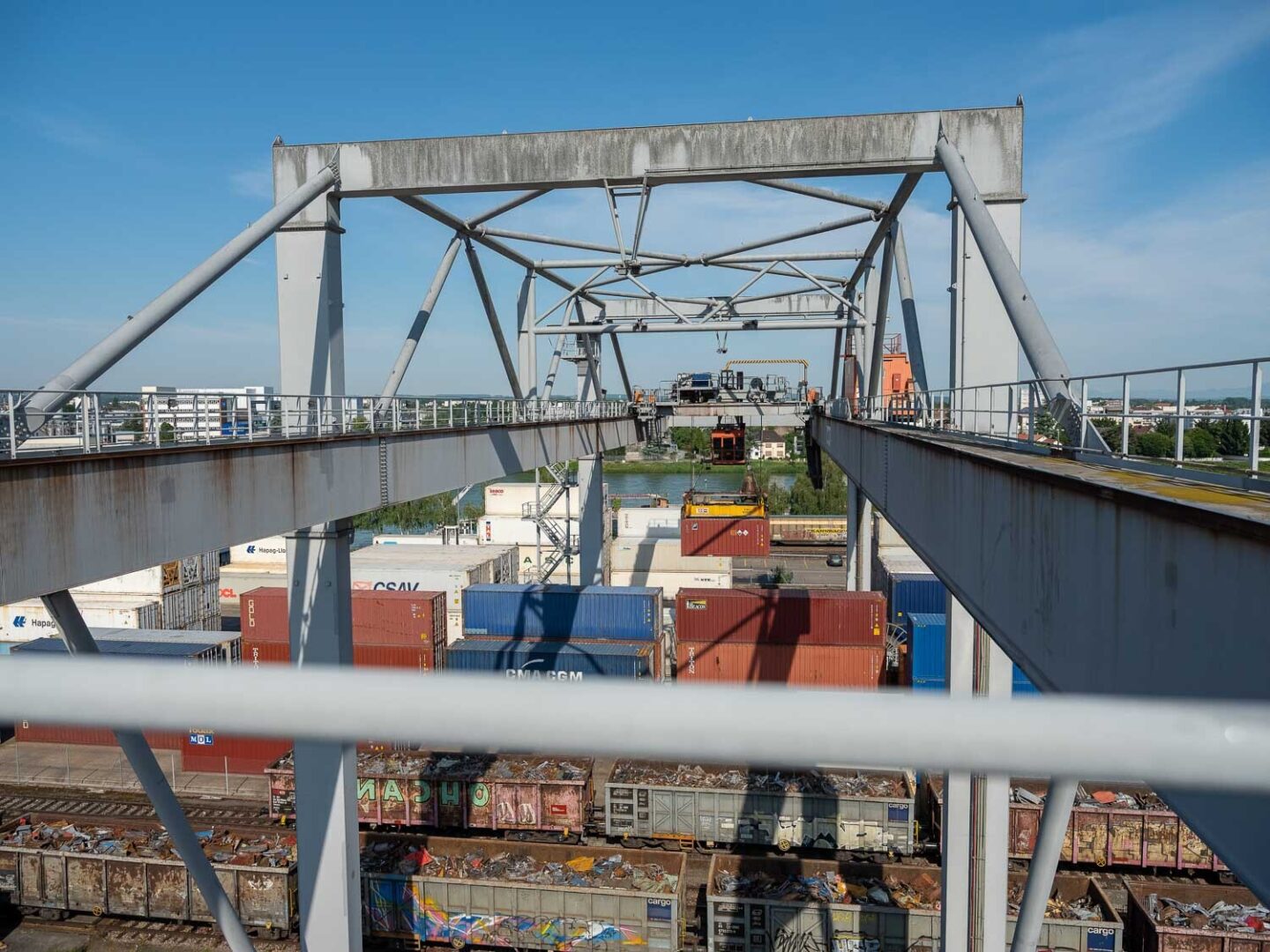 A large metal gantry crane overlooks a shipping container yard and a body of water.