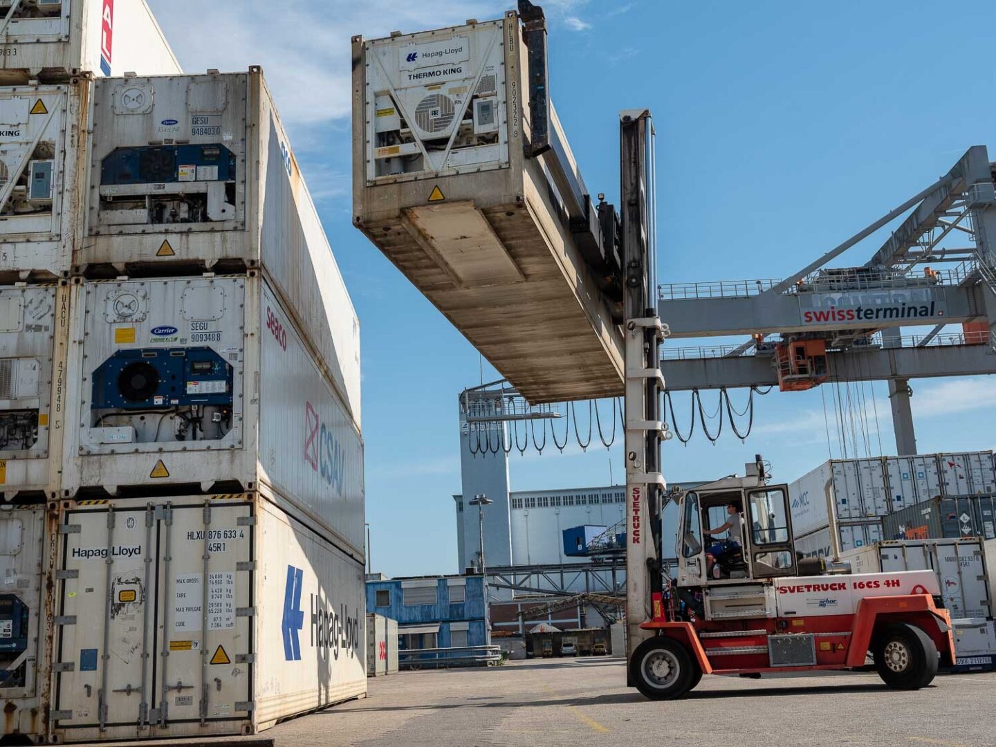A forklift moves shipping containers at a port with a large ship in the background.