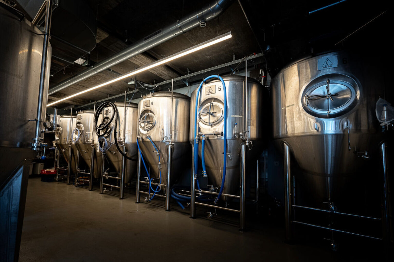A row of large, shiny stainless steel brewing tanks in a dimly lit room.
