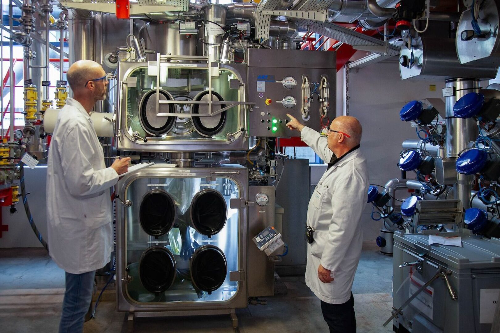 Zwei Männer in Laborkitteln diskutieren eine Maschine mit mehreren runden Behältern.