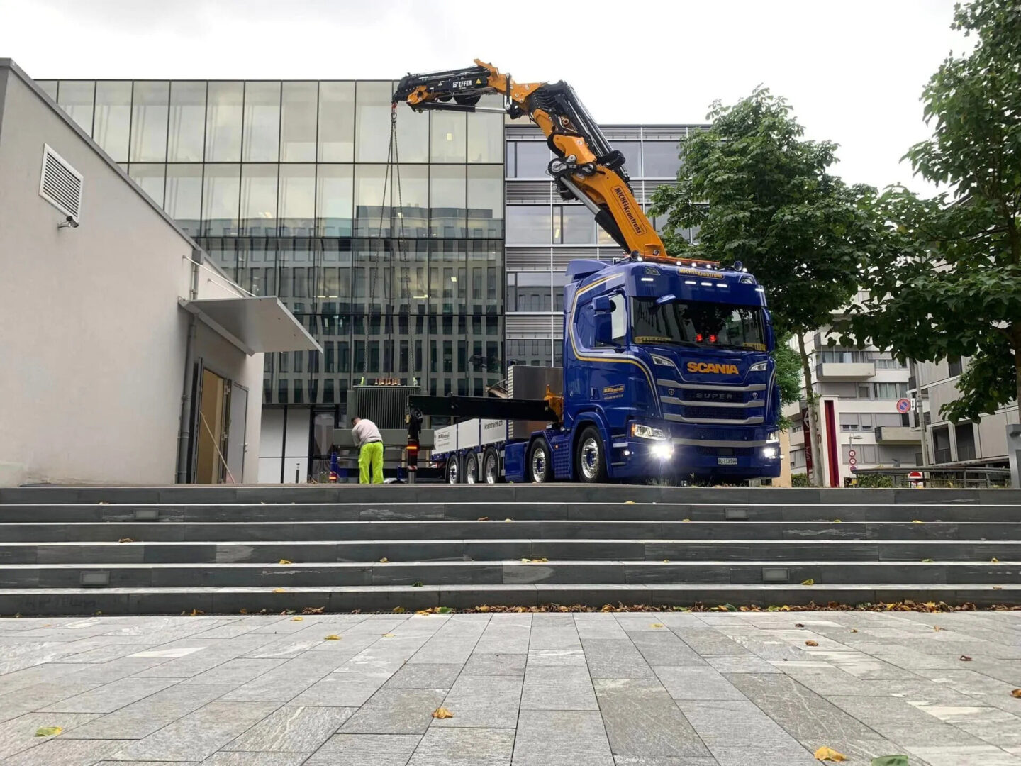 Ein blauer Scania-Lastwagen mit einem Kran hebt etwas vor einem Gebäude an.