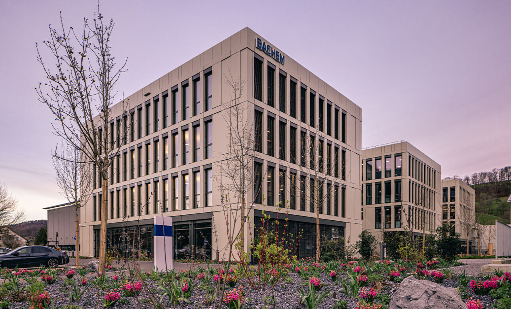 Moderne Bürogebäude mit mehreren Stockwerken und vielen Fenstern, umgeben von Blumenbeeten.
