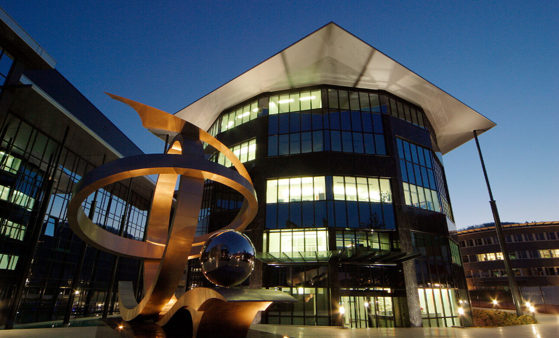 Moderne Glasgebäude mit Skulptur davor bei Nacht.