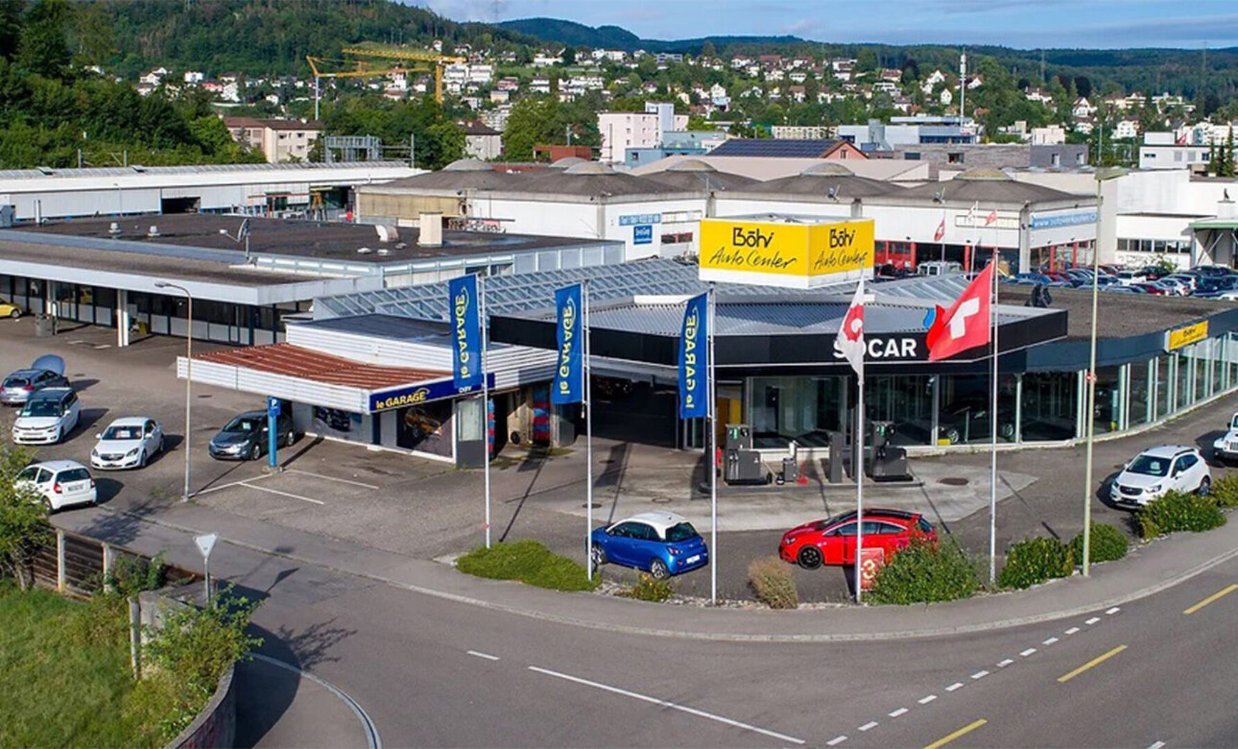 Autohaus mit mehreren Ausstellungsfahrzeugen und Schweizer Flaggen vor einem Gebäude.