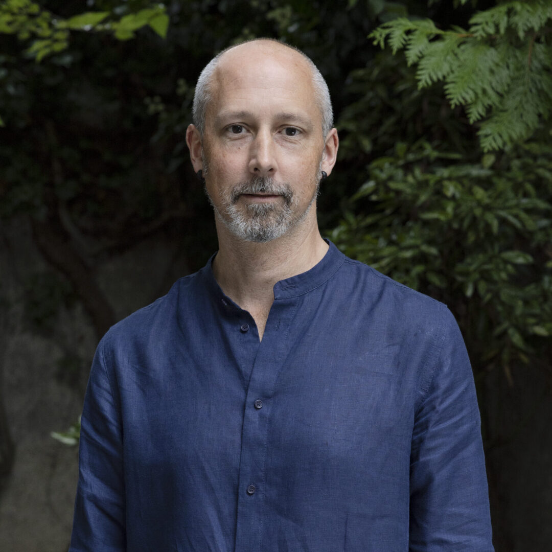 Mann mit grauem Bart und glatzer Kopf trägt blaues Hemd vor grünem Hintergrund.