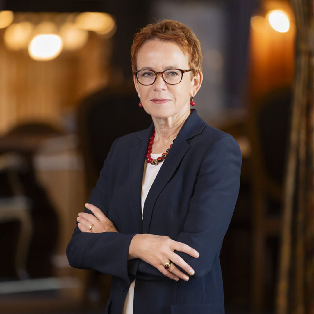 Eine Frau mit Brille steht mit verschränkten Armen in einem Raum mit warmem Licht.