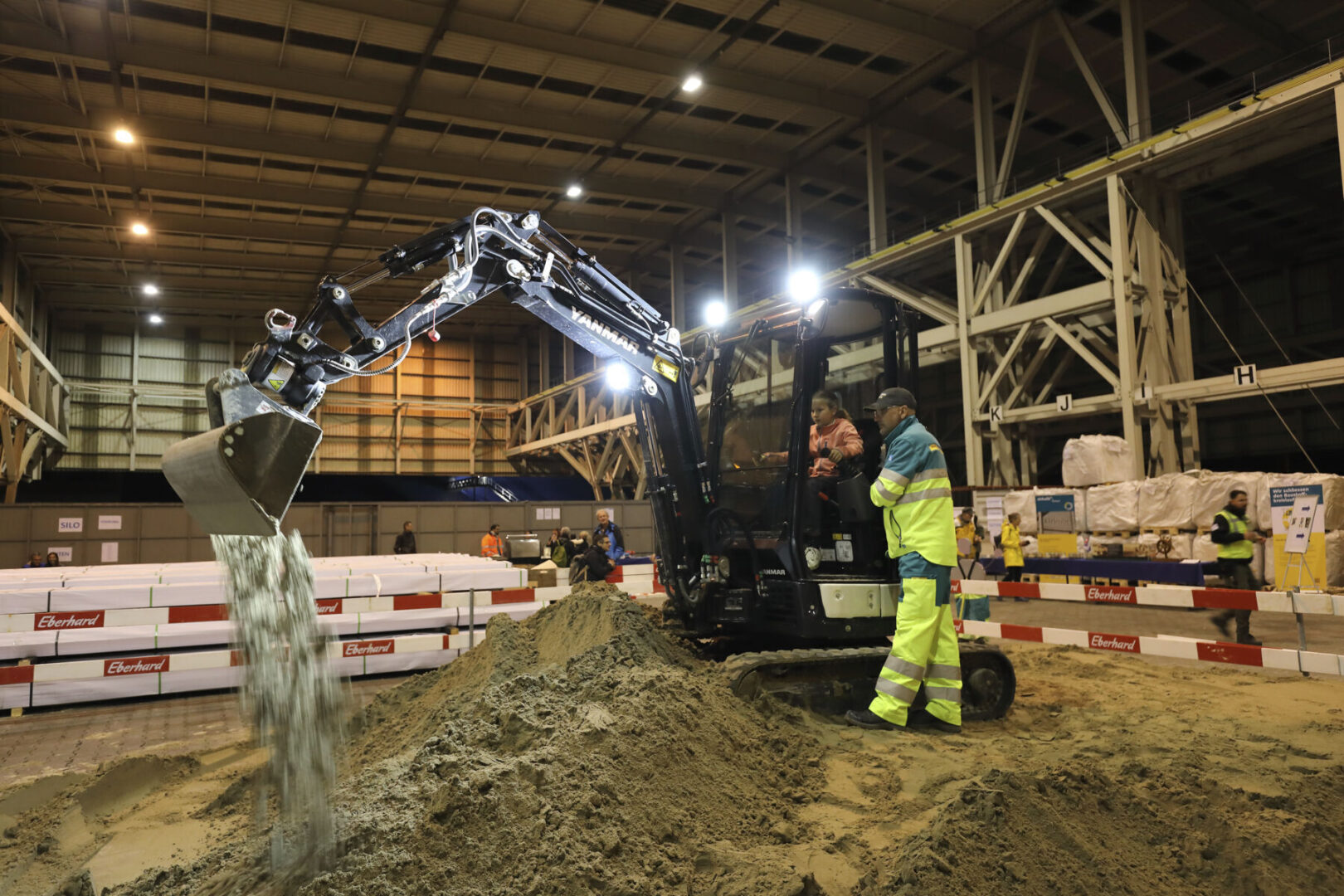 Ein Bagger gießt Sand in einer Lagerhalle, während Arbeiter zusehen.