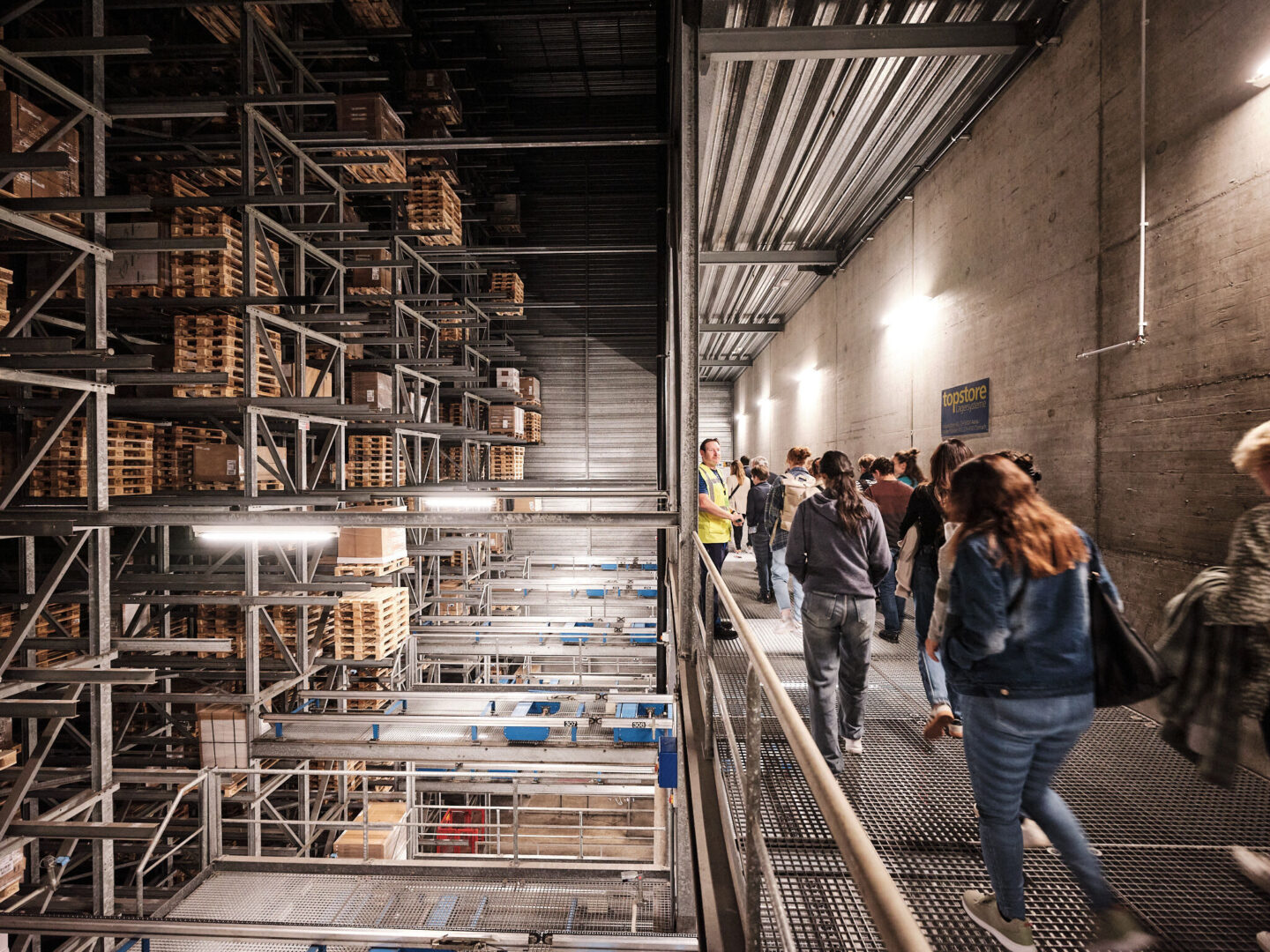 Eine Gruppe von Menschen geht durch ein Lagerhaus mit hohen Regalen voller Paletten.