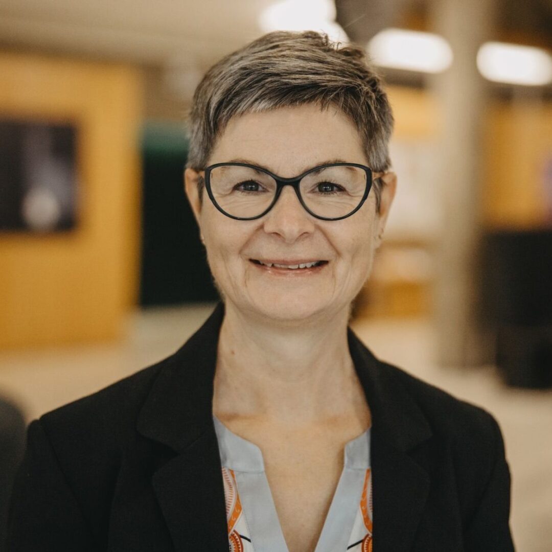 Eine lächelnde Frau mit kurzen Haaren und Brille in einem schwarzen Blazer.