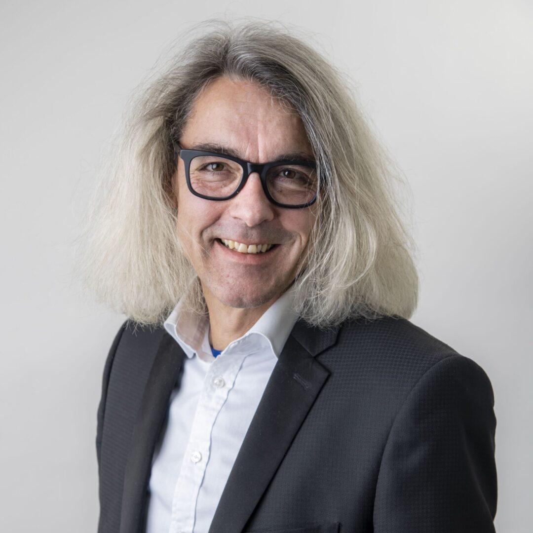 Ein lächelnder Mann mit grauen Haaren und Brille trägt ein weißes Hemd und einen dunklen Anzug.