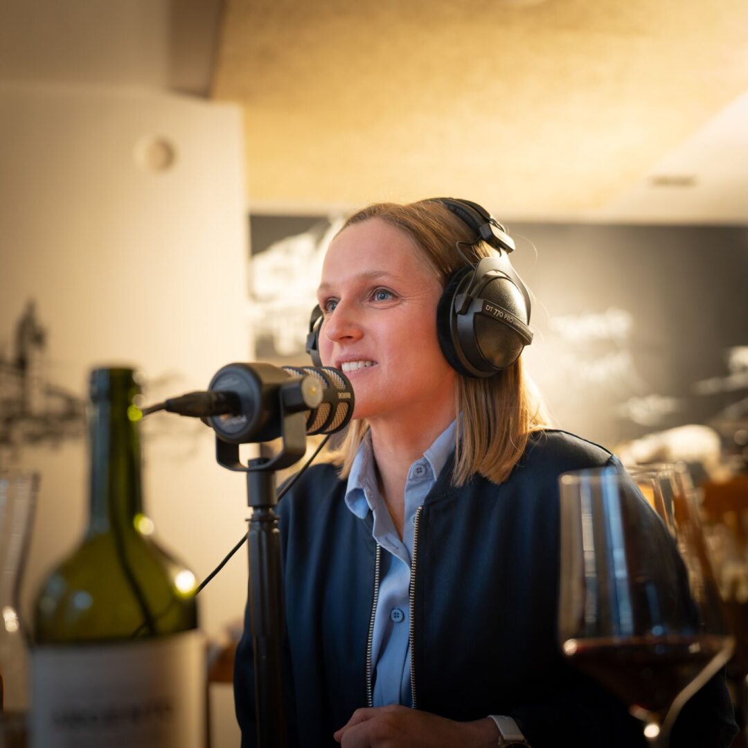 Eine Frau mit Kopfhörern spricht in ein Mikrofon vor Weinflaschen.